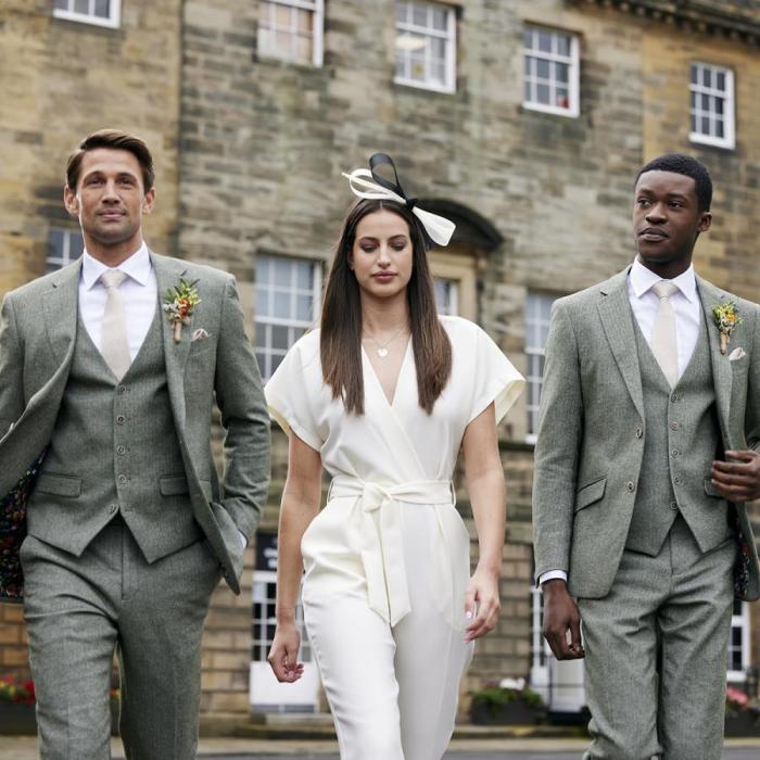 Two men in smart grey wedding suits and woman in a cream jumpsuit and fascinator